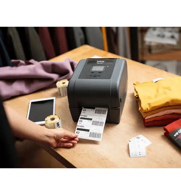 Imprimante de bureau Brother TD4650 imprimant des étiquettes, avec tablette et vêtements à proximité.