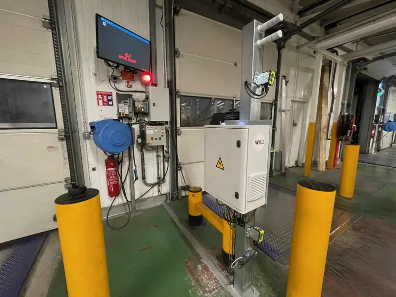 Le quai de chargement d'un entrepôt comporte des équipements de sécurité, des panneaux de contrôle, un extincteur, un grand écran affichant "pas de signal" et des bornes jaunes entourant un poste de pesage ou de scannage automatisé.