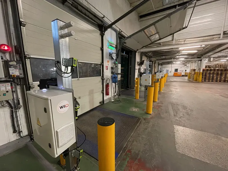 Quai de chargement d'un entrepôt avec des portes ouvertes, des bornes de protection jaunes, des panneaux de contrôle et des équipements électroniques. Des palettes sont empilées à l'arrière-plan sous un éclairage fluorescent.