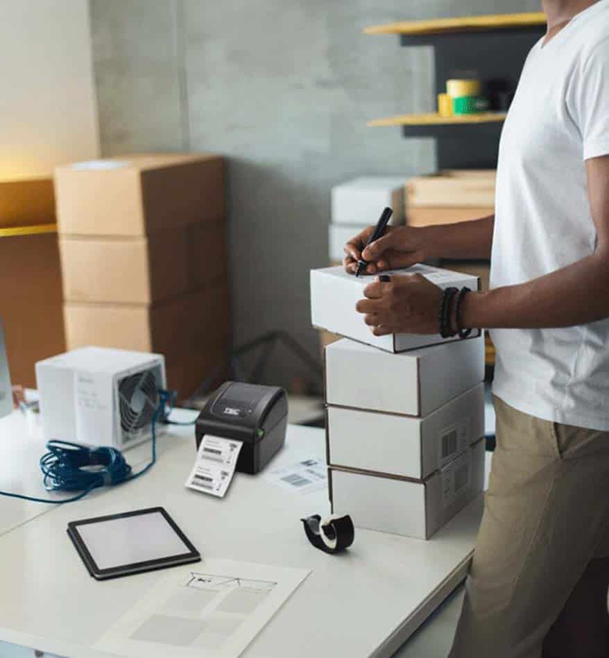 Homme préparant des colis avec une imprimante de bureau TSC DA210/220. Boîtes, étiquettes et tablette visibles.