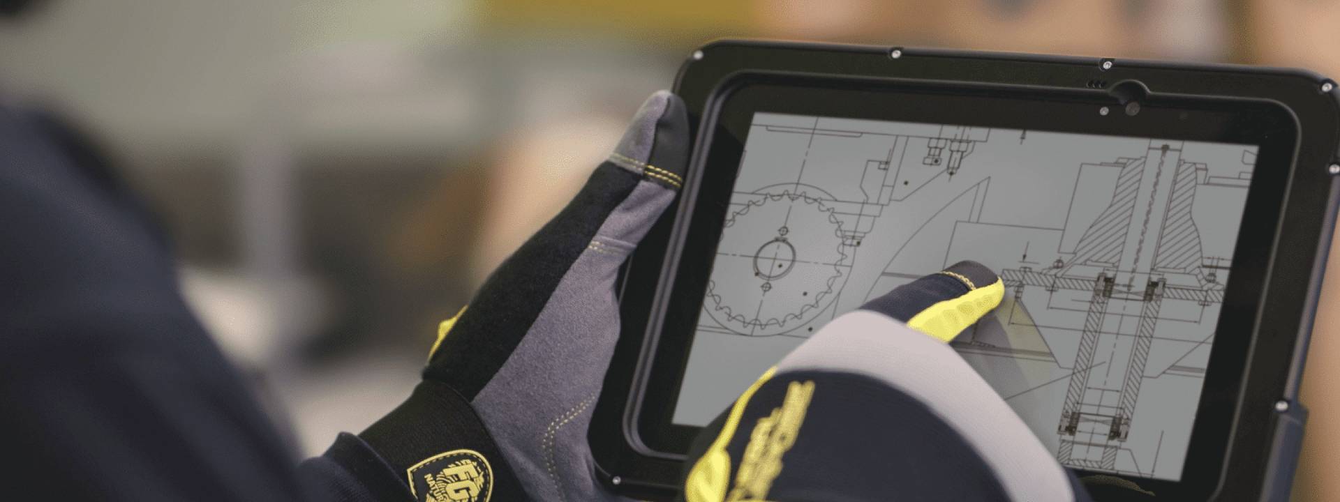 Une personne portant des gants de protection utilise une tablette affichant un dessin technique détaillé avec des engrenages, des composants de machines et du matériel connexe.